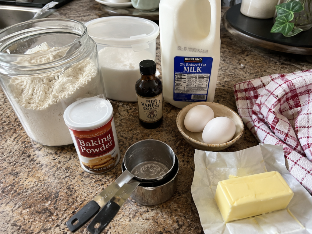 Fresh pancake ingredients including flour, eggs, milk, butter, vanilla extract, sugar, and baking powder arranged on a kitchen countertop.